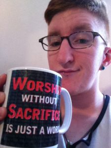 Taylor on Set of First Porn Shoot. They are smirking and holding a mug that says "Worship without Sacrifice is just a word"
