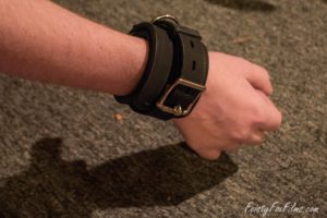 Taylor's right arm is extended, palm facing away from the camera. They are wearing Stockroom's thick silicone bondage cuff tightly around their wrist.