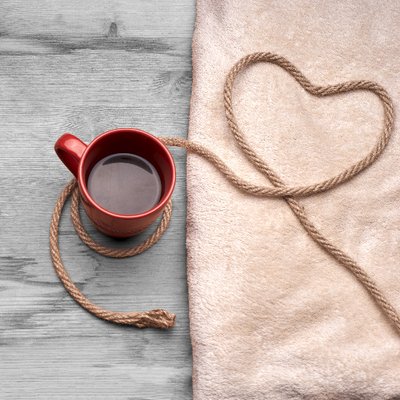 Coffee and Kink's logo; a full, red coffee mug sits on light grey wood. next to it there is tan, soft-looking fabric. On top of the fabric there is some undyed rope on top of the fabric that is bent int a heart shape before forming a loop around the coffee mug.