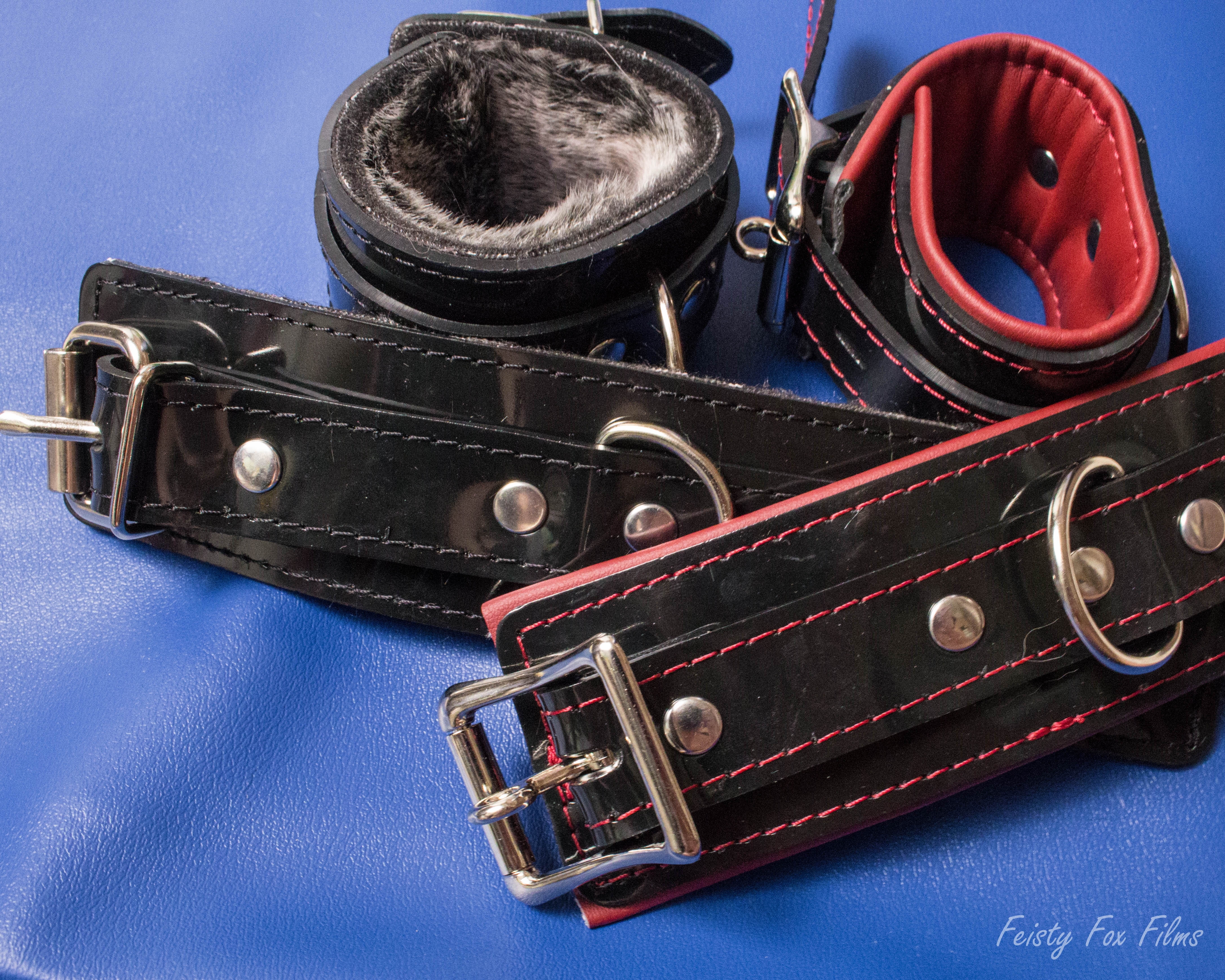 The Fur Lined Cuffs and Padded Lockable Cuffs sit on top of a sheet of blue vinyl. In the back, one of each cuff is closed and sitting up. In front, one of each cuff rests open against its match.