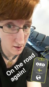 A very sleepy Taylor has a quasi-surprised look on their face. Text on the image says "on the road again!" and there's a logo indicating they're in an airport in Chicago. You can see a row of airport chairs over their shoulder.