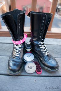 Tall black Doc Martins are sitting mostly unlaced on a deck. They are shiny, though a bit scuffed up, and their laces are white. Between the two shoes are a small pile of boot blacking materials. Attached around the ankle of one of the boots is a light pink collar with white dog bones on it.