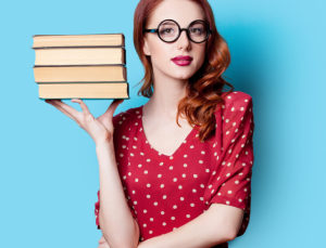 A very pretty redhead stands holding four books by her head. Her hair is just past her shoulders and wavy, pulled to one side. She is wearing large framed blackglasses, red lipstick, and a red dress with white polka dots.
