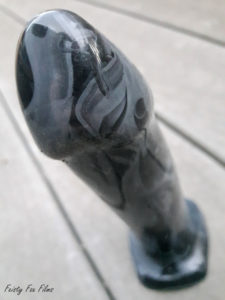 Servant Sex Toy's Lune stands on a deck,tip pointed toward the camera. The base is out of focus, the head and top portion of the shaft in focus, highlighting how the black and grey silicone blends together in a striking and beautiful manner.