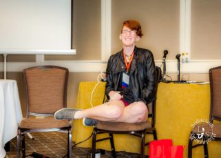 Taylor sits on a dining chair holding a microphone and grinning widely. Their legs are tucked underneath them, legs sticking out the side, and they're wearing sneakers, short red shorts, a fitted black tank top, and a worn black leather jacket. Their hair is swooping to one side and they're wearing a Woodhull name badge.
