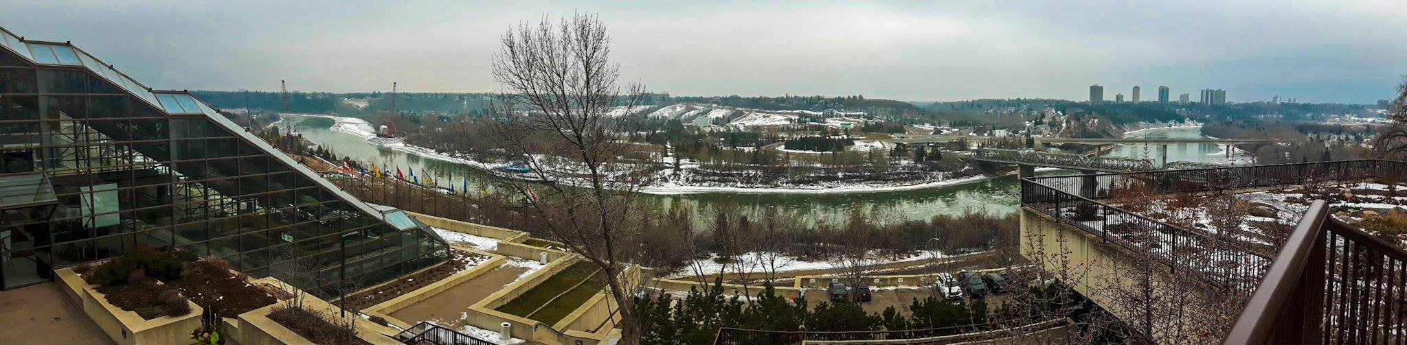 panorama of a riverfront. it's winter and there is snow everywhere, the river partly iced over. the greens, blues, and teals in the water and overcast sky are very vibrant. in the forefront of the image there are some buildings, steps, and little planters. there are bridges crossing the river at each end