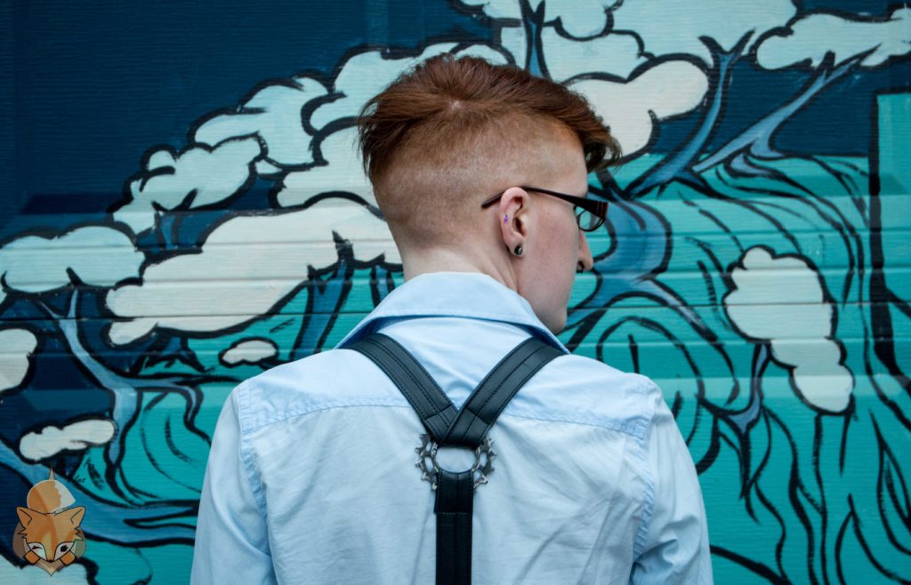 Taylor stands facing a garage door with some sort of blue and green painting on it. They're wearing a light blue button-up shirt, black bike tube suspenders with a metal gear in the center. Their hair is shaved on the sides and back, a bit longer on the top.