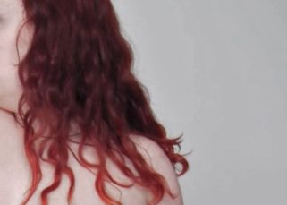 The right side of a pale white person with died red hair is shown from the chest up. They are topless and their face is blurred out.